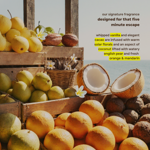 Fruit display with pears, oranges, and coconuts by a body of water, with text about a fragrance.